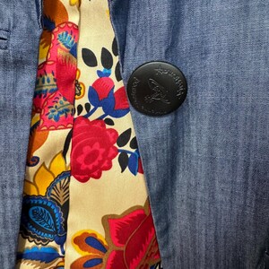 May include: A blue denim blazer featuring a vibrant floral lining with red, yellow, and blue patterns. A large, round black button is attached to the front, embossed with the text Vivienne Westwood and a small orb logo design in the center.