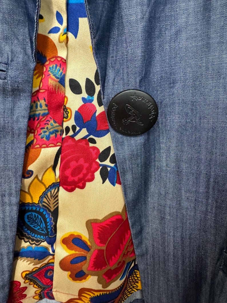 May include: A blue denim blazer featuring a vibrant floral lining with red, yellow, and blue patterns. A large, round black button is attached to the front, embossed with the text Vivienne Westwood and a small orb logo design in the center.