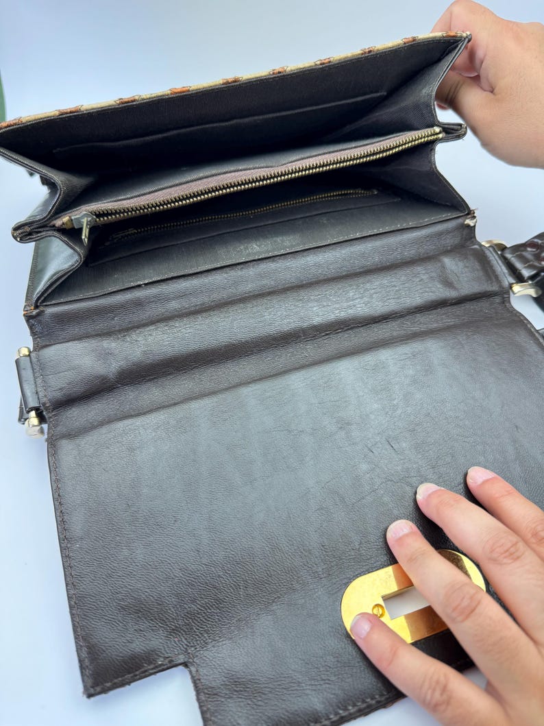 May include: Open view of a brown leather handbag with a gold-colored clasp. The interior features a zippered pocket and multiple compartments. The exterior has a patterned design on the flap.