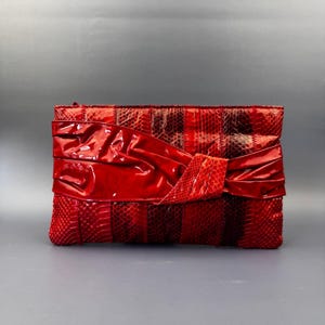 Puede incluir: Un bolso de mano rectangular con una textura de piel de serpiente en tonos rojo y rojo oscuro. La parte frontal presenta una banda horizontal ancha de material rojo brillante y charol, fruncida y anudada en el lado derecho del bolso.