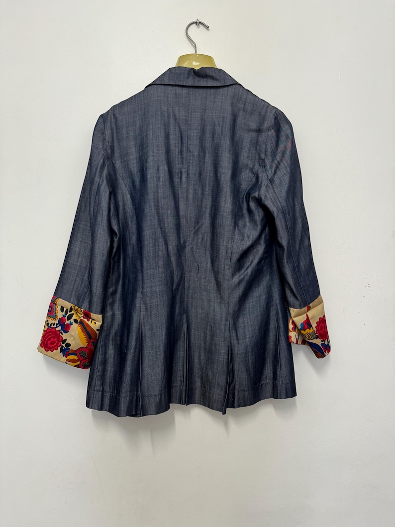 May include: A dark blue blazer with a notched collar and long sleeves. The sleeve cuffs are turned up to reveal a beige lining featuring a vibrant, multi-colored floral pattern in shades of red, blue, yellow, and green against a cream background.
