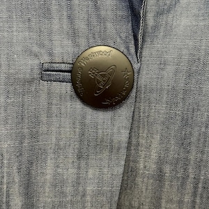 May include: A blue denim blazer featuring a large, circular, matte black button. The button is embossed with the text Vivienne Westwood and a central orb logo. The fabric has a visible, fine-grained texture with subtle vertical variations in color.