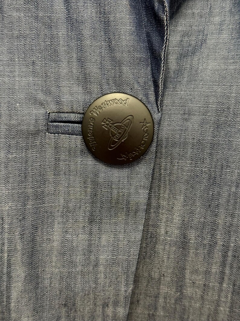 May include: A blue denim blazer featuring a large, circular, matte black button. The button is embossed with the text Vivienne Westwood and a central orb logo. The fabric has a visible, fine-grained texture with subtle vertical variations in color.