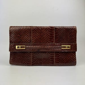 Könnte beinhalten: Eine rechteckige braune Clutch mit texturiertem Schlangenledermuster. Die Frontklappe wird durch einen horizontalen Riemen gesichert, der mit zwei rechteckigen Akzenten aus poliertem goldfarbenem Metall verziert ist und ein strukturiertes Aussehen bietet.