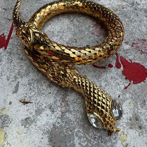 Op de afbeelding: Een goudkleurige slangvormige armband met een textuur, schubachtig ontwerp. De kop van de slang heeft twee heldere, druppelvormige edelstenen als ogen. De armband is opgerold, waarbij de kop en staart elkaar bijna raken.