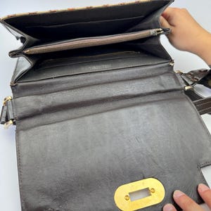 May include: Open brown leather handbag with a gold-colored clasp. The interior features multiple compartments, including a zippered pocket. The top flap has a patterned design in shades of brown and tan. The bag has a shoulder strap.