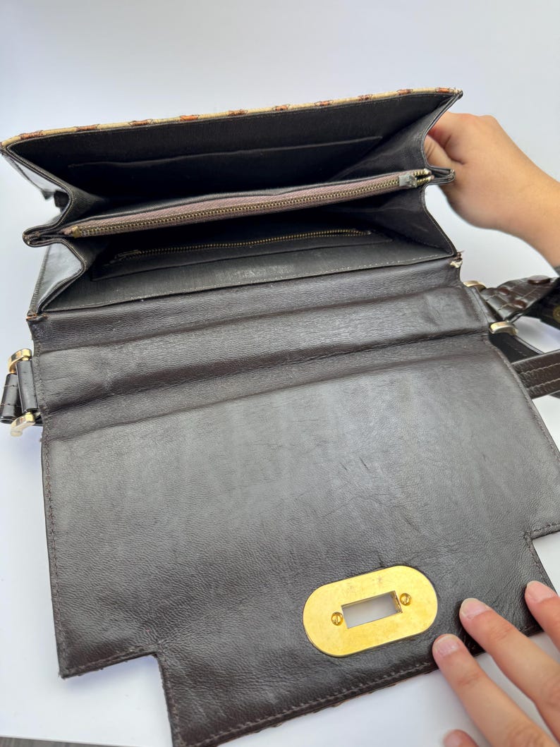 May include: Open brown leather handbag with a gold-colored clasp. The interior features multiple compartments, including a zippered pocket. The top flap has a patterned design in shades of brown and tan. The bag has a shoulder strap.