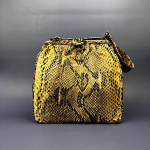 Könnte beinhalten: Eine strukturierte Handtasche mit einem leuchtend gelben und schwarzen Schlangenledermuster. Die Tasche hat eine rechteckige Form mit abgerundeten Ecken, einen Rahmenverschluss mit zentralem Metallverschluss und einen kurzen Tragegriff.