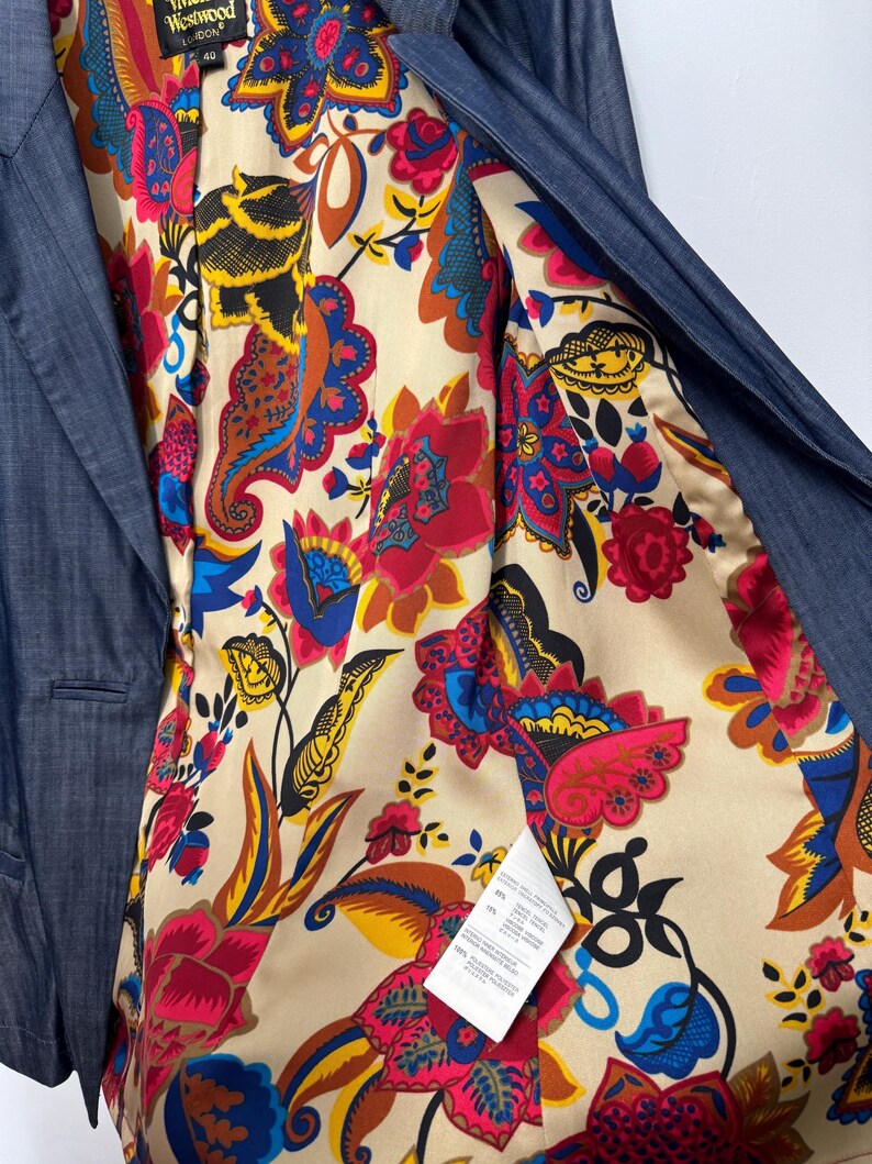 May include: A dark blue blazer with a vibrant, multicolored floral and paisley print lining. The interior features a Vivienne Westwood London label and a white care tag. The fabric has a subtle texture, and the lining displays bold red, yellow, and blue patterns on a cream background.