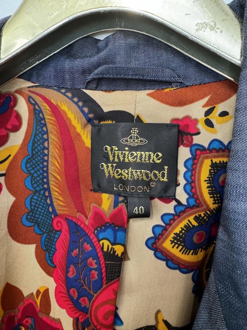 May include: A blue denim jacket featuring a vibrant, multicolored paisley print lining in shades of red, yellow, and blue. A black fabric label is stitched inside, displaying the text Vivienne Westwood London and the number 40.