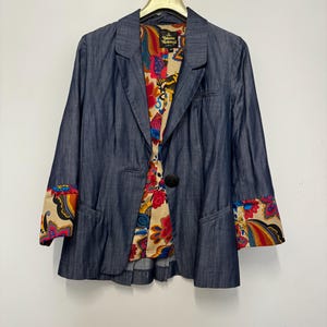 May include: A blue denim blazer featuring a single black button closure, notched lapels, and long sleeves with cuffs that reveal a vibrant, multicolored floral and paisley pattern lining, which also lines the interior of the garment.