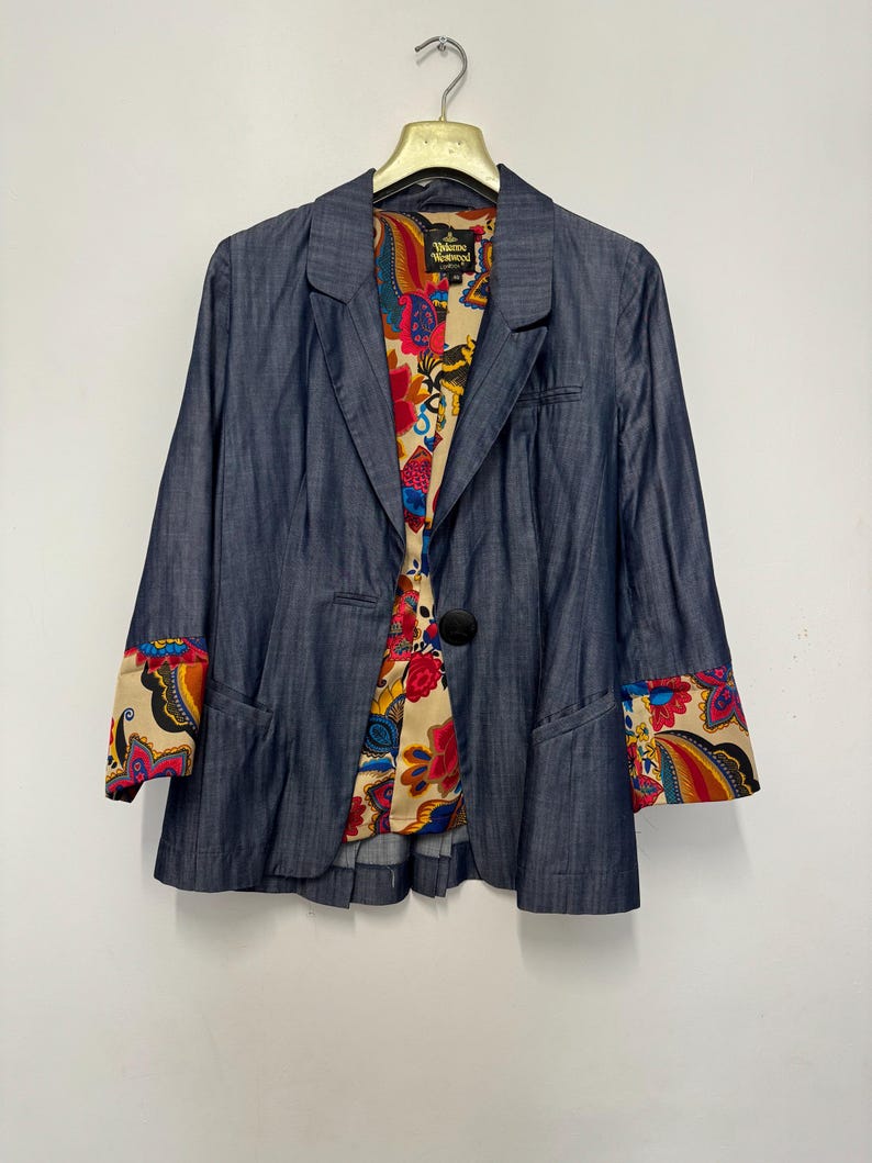 May include: A blue denim blazer featuring a single black button closure, notched lapels, and long sleeves with cuffs that reveal a vibrant, multicolored floral and paisley pattern lining, which also lines the interior of the garment.