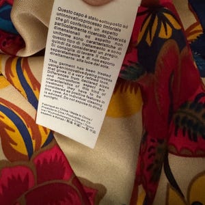 May include: A white fabric care label attached to a garment with a colorful floral pattern in shades of red, yellow, and blue. The label contains printed text in Italian, English, and other languages regarding a specialized dyeing process.