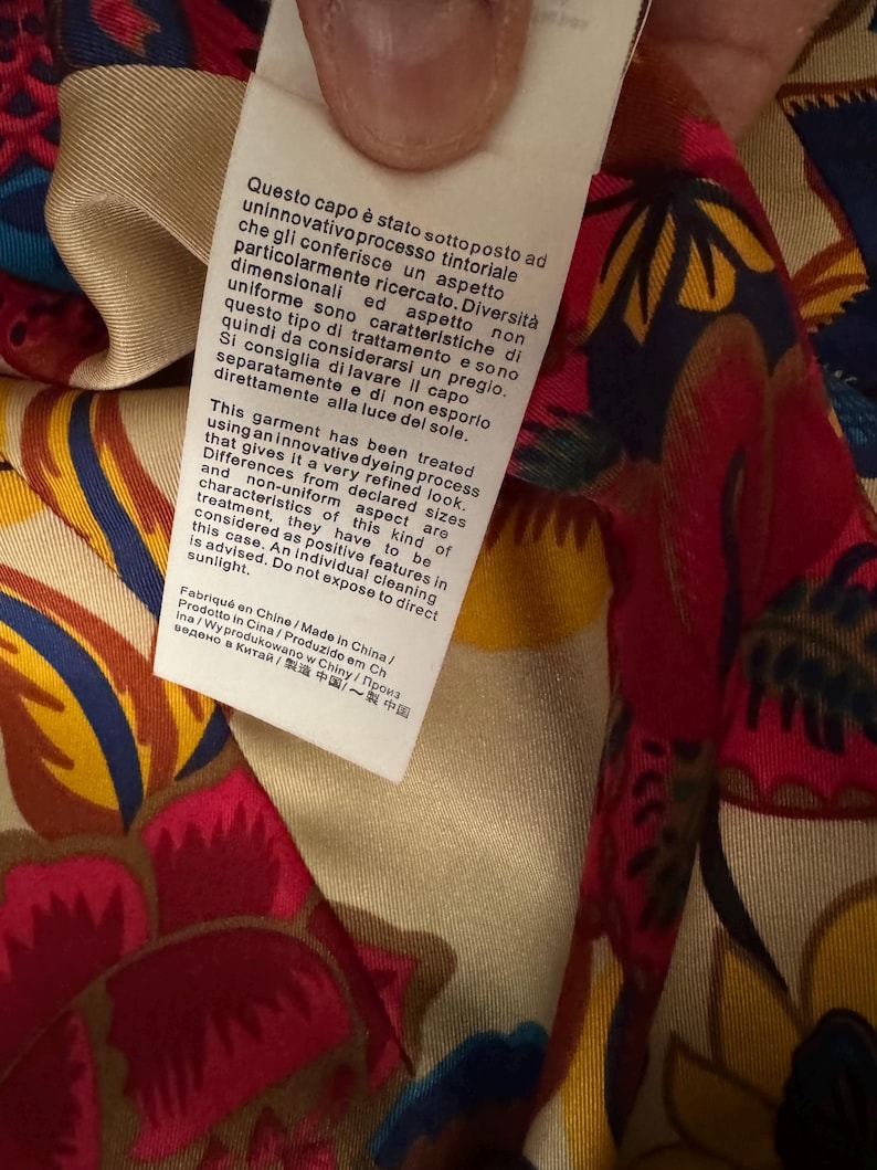May include: A white fabric care label attached to a garment with a colorful floral pattern in shades of red, yellow, and blue. The label contains printed text in Italian, English, and other languages regarding a specialized dyeing process.