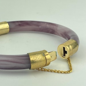 Może przedstawiać: Otwierana bransoletka typu bangle z półprzezroczystą, marmurkową opaską w kolorze lawendowym i białym. Bransoletka posiada złote metalowe końcówki oraz bezpieczny mechanizm zamykający, uzupełniony delikatnym złotym łańcuszkiem zabezpieczającym.