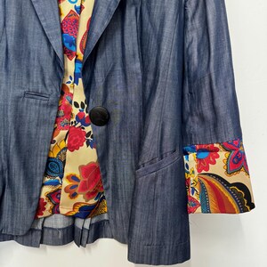May include: A blue blazer with a single black button closure, featuring a cream-colored lining and sleeve cuffs decorated with a vibrant, multi-colored floral and paisley pattern in shades of red, yellow, blue, and black.
