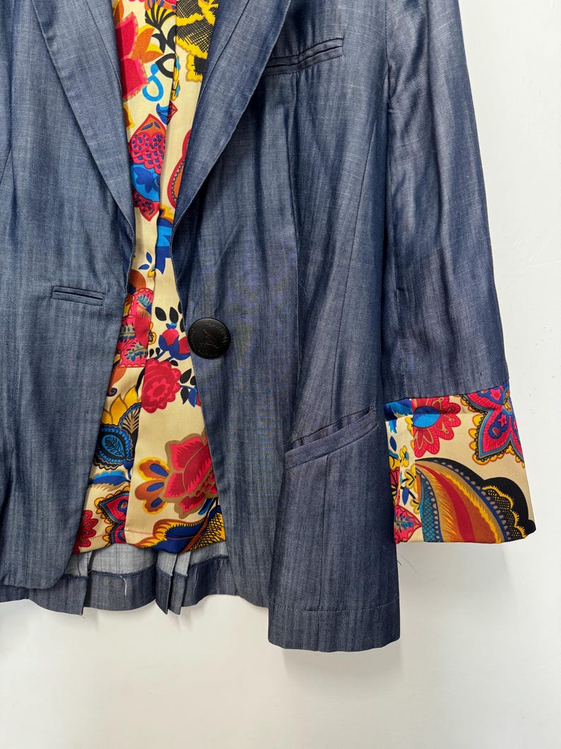 May include: A blue blazer with a single black button closure, featuring a cream-colored lining and sleeve cuffs decorated with a vibrant, multi-colored floral and paisley pattern in shades of red, yellow, blue, and black.
