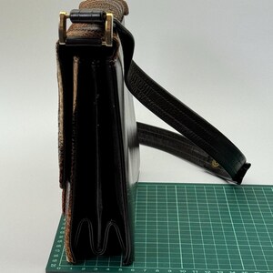 May include: A black leather satchel with a textured flap and a gold-colored clasp. The bag has a long, dark brown shoulder strap. The satchel is approximately 10 inches wide.