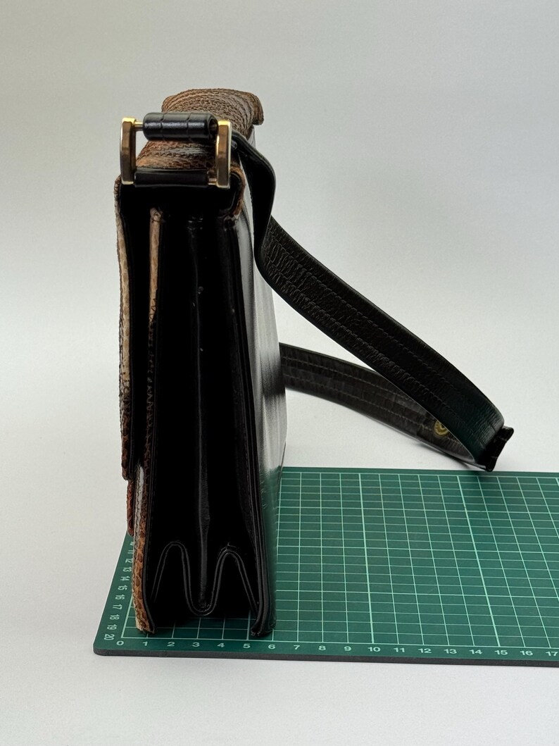 May include: A black leather satchel with a textured flap and a gold-colored clasp. The bag has a long, dark brown shoulder strap. The satchel is approximately 10 inches wide.