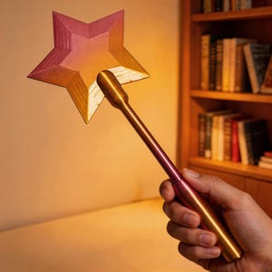 May include: A star-shaped wand with a metallic finish. The star is a gradient of pink and gold, and the handle is a matching gold and pink. The wand is held in a hand, with a bookshelf in the background.