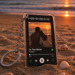 Peut inclure: Un porte-clés en acrylique transparent avec une chaîne et un anneau argentés. Le porte-clés présente une photo d'un couple au coucher du soleil et le texte "To The Moon" avec une interface de lecteur de musique. Le fond est une plage de sable.