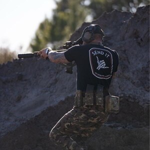May include: A person wearing a black t-shirt with the text "SEND IT" and a hand gesture graphic, camouflage pants, and a tactical belt, aiming a rifle. The person is outdoors, on a dirt mound.