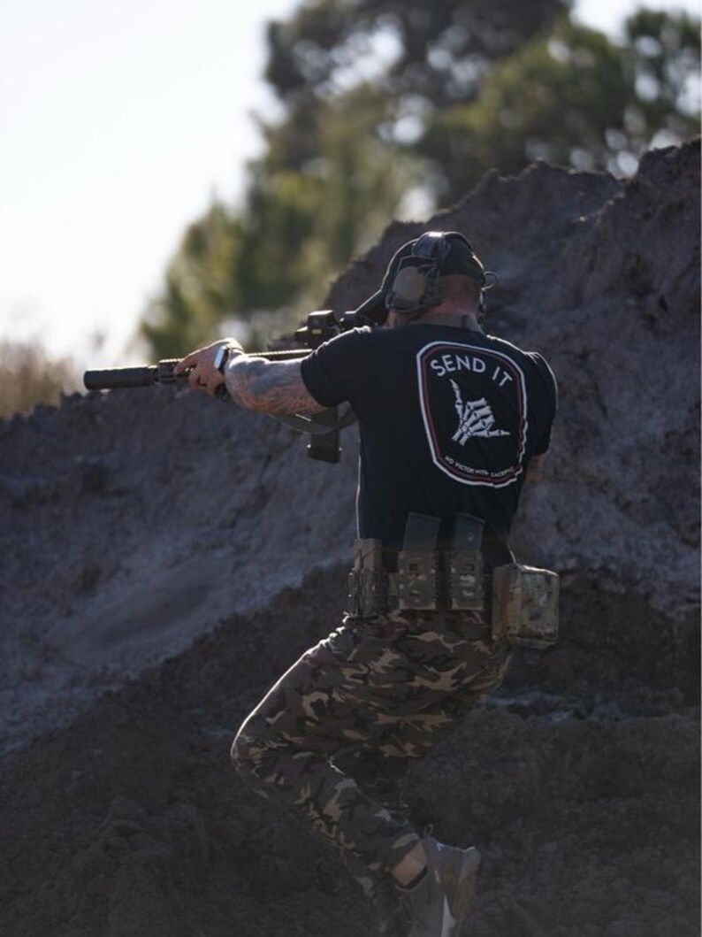 May include: A person wearing a black t-shirt with the text "SEND IT" and a hand gesture graphic, camouflage pants, and a tactical belt, aiming a rifle. The person is outdoors, on a dirt mound.