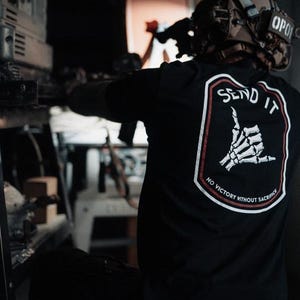 May include: Black t-shirt with a white and red graphic on the back. The graphic features a skeleton hand in a shaka sign with the words "SEND IT" and "NO VICTORY WITHOUT SACRIFICE". The person is wearing a helmet.