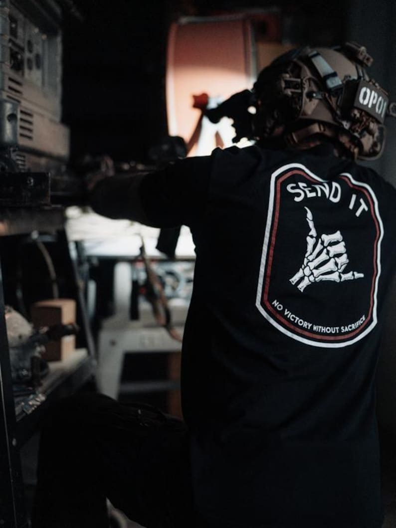 May include: Black t-shirt with a white and red graphic on the back. The graphic features a skeleton hand in a shaka sign with the words "SEND IT" and "NO VICTORY WITHOUT SACRIFICE". The person is wearing a helmet.