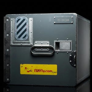 Pode incluir: Uma caixa de metal cinza com formato retangular, com uma alça, uma trava e um rótulo com listras diagonais. A caixa tem um adesivo amarelo com o texto "rUIfly.com". A caixa tem o número "03 D0288".