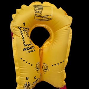 Könnte beinhalten: Hellgelbe Schwimmweste für Erwachsene/Kinder mit schwarzen Anweisungsdiagrammen und Text. Die Schwimmweste hat ein kreisförmiges Design mit einer Halsöffnung und einem Schnallenverschluss. Die Weste hat ein Etikett mit Text.