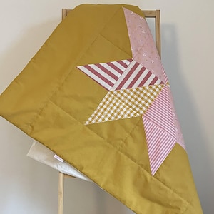 Può includere: Una trapunta fatta a mano con un motivo a stella. La trapunta presenta un bordo e un retro giallo senape, con una stella patchwork nei toni del rosa, bianco e giallo. Il design a stella include motivi a righe e a quadretti.
