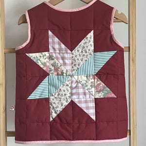 Könnte beinhalten: Gesteppte Weste in tiefem Burgunderrot mit einem großen Sternmuster in der Mitte. Der Stern besteht aus verschiedenen gemusterten Stoffen, darunter Blumen-, Streifen- und Vichymuster. Die Weste hat eine rosa Einfassung.