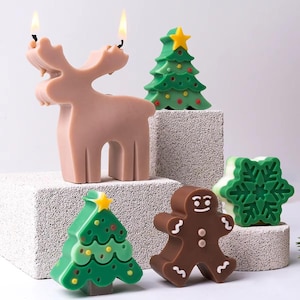 Può includere: Una collezione di candele in cera a tema natalizio che include una renna marrone chiaro con due stoppini accesi, due alberi di Natale verdi con stelle gialle e decorazioni colorate, un omino di pan di zenzero e una candela a forma di fiocco di neve.