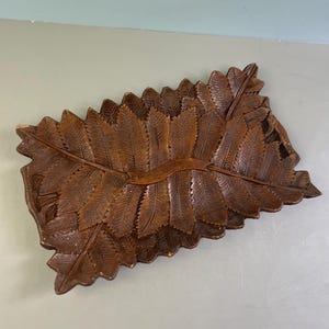 May include: A rectangular wooden tray featuring a detailed carved pattern of overlapping fern leaves. The dark brown wood surface displays intricate textures and veins on each leaf, with a central horizontal band crossing the design.