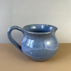 May include: A small, round, blue ceramic pitcher with a handle and a pouring spout. The pitcher has a speckled texture and a glossy finish. The handle is curved and attached to the side of the pitcher. The spout is slightly angled.