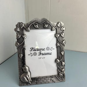 May include: A silver-colored picture frame with a cat design. The frame has a brick-like border and is adorned with leaf and vine accents. The text "Picture Frame" is displayed, with dimensions of 3.5" x 5".