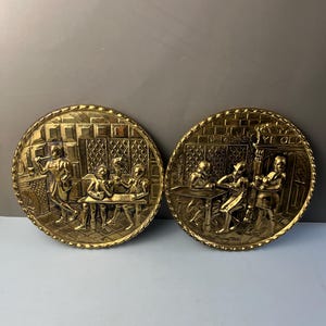 May include: Two circular brass wall plaques featuring embossed tavern scenes. The left plaque depicts figures at a table and a fireplace. The right plaque shows figures at a table with the text YE OLD inscribed above an arched doorway.