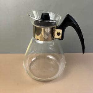 May include: A clear glass coffee carafe featuring a wide, rounded base and a tapered neck. It includes a black plastic lid and a matching black ergonomic handle attached to a polished metallic band that encircles the upper portion of the vessel.