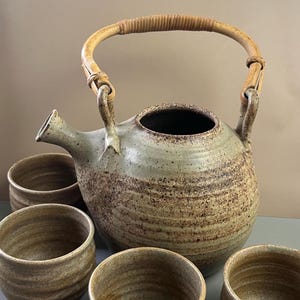 May include: A ceramic teapot with a speckled light green and brown glaze, featuring a curved spout and a rounded body. The teapot has a light-colored, arched bamboo handle attached to two ceramic loops. Four matching ceramic cups are arranged below.