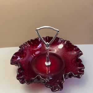 May include: A ruby red, two-tiered serving dish with a ruffled edge and a silver handle. The dish has a textured surface and a circular base. The handle is a simple, arched design. The dish is likely made of glass.