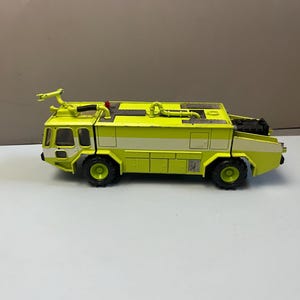 May include: A lime green and white toy fire truck featuring a roof-mounted water cannon, a small red light, black textured wheels, and a flatbed design with visible piping details along the top surface of the vehicle body.