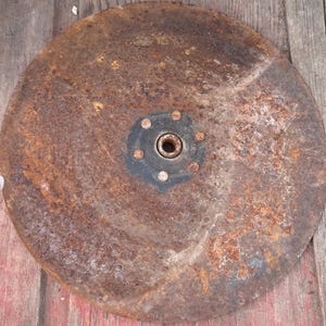 May include: A circular, rusty metal disc with a central hub and bolt pattern. The disc has a weathered, aged appearance with a mix of brown and orange hues, suggesting exposure to the elements. The central hub is dark with a circular opening.