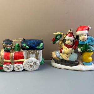 May include: Two Christmas ornaments: a colorful train and a figurine of two children sledding. The train is red, white, and gold with a blue roof. The children are wearing winter clothes, including red hats and scarves.