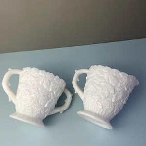 May include: Two white milk glass cream pitchers with a raised leaf design. Each pitcher has a handle and a spout. The pitchers are on a light blue surface.