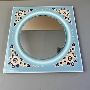 May include: Square, light blue mirror with a round center. The frame features a floral design in cream and navy blue at each corner. The mirror is a decorative item.