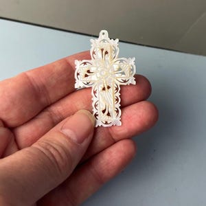 May include: A white mother-of-pearl cross pendant featuring intricate, delicate cut-out filigree patterns throughout the surface. The cross has a small circular hole at the top for attachment and displays a natural iridescent sheen across its carved surface.