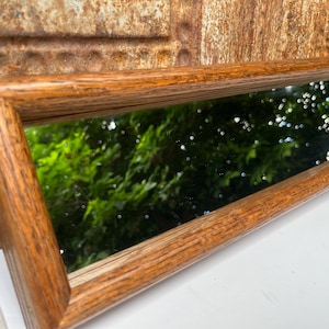 May include: A long, rectangular mirror featuring a thick, natural-toned wooden frame with a visible grain pattern. The reflective glass surface shows a clear, angled view of green foliage and dappled sunlight, set within a sturdy, mitered wood border.