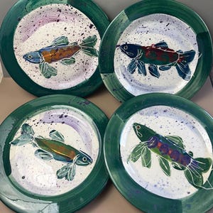 May include: Four ceramic plates with hand-painted fish designs. Each plate features a white center with a colorful fish illustration, surrounded by a green border. The fish are in shades of blue, orange, green, and purple, with white outlines.