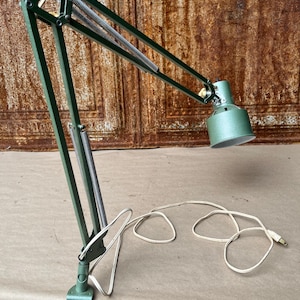 May include: A green metal architect lamp with an adjustable articulated arm, exposed silver springs, and a clamp base. The lamp features a cylindrical shade and includes a white electrical cord with a standard two-prong plug for power connection.